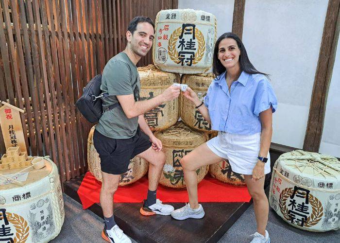 Two smiling guests of a sake museum tour and tasting in Fushimi, raising sake cups in front of sake barrels.